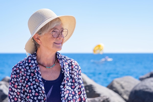 Attraktive Seniorin mit Hut und Brille, die am Meer sitzt und die Schönheit in der Natur bewundert. Lächelnde kaukasische Frau, die einen sonnigen und sommerlichen Tag im Ruhestand oder Urlaub genießt