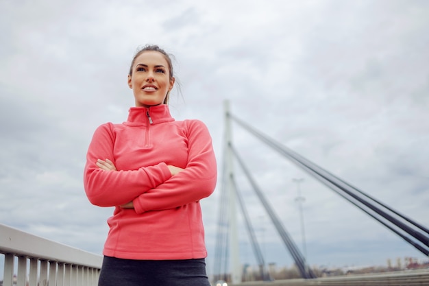 Attraktive Sportlerin in ausgezeichneter Form, die mit verschränkten Armen auf der Brücke steht und stolz ist, weil sie eine Leistung erbracht hat.