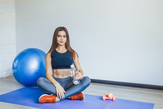 Attraktive sportliche Frau mit blauem Ball, der auf der Matte sitzt und zur Kamera schaut.