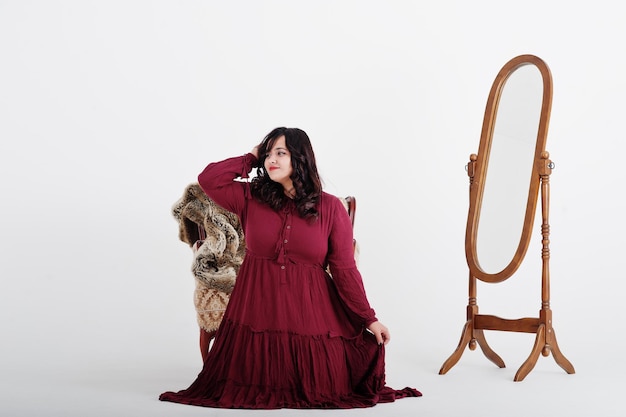 Attraktive südasiatische Frau in tiefrotem Kleid posiert im Studio auf weißem Hintergrund vor Spiegel und Stuhl