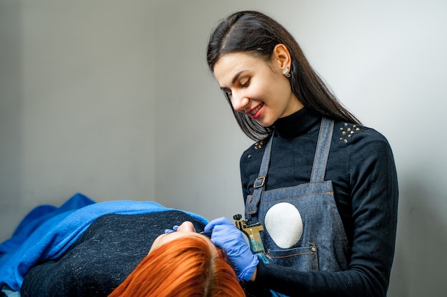 Attraktive Tätowiererfrau mit dem langen Haar, das dauerhaftes Make-up macht und zu einem Patienten im Badekurortsalon lächelt.