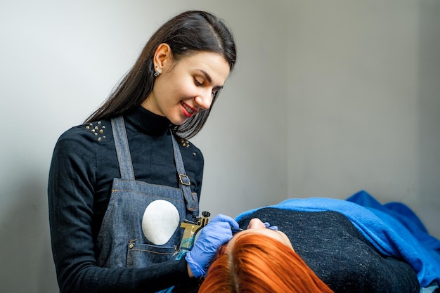 Attraktive Tätowiererin mit langen Haaren, die Permanent Make-up macht und einem Patienten im Spa-Salon zulächelt Schöne rothaarige Frau, die sich auf den Lippen tätowieren lässt