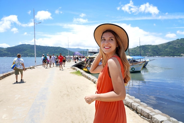 Attraktive Touristenfrau am Pier dreht sich um und schaut lächelnd weg, bereit für eine Bootsfahrt in einem tropischen Reiseziel