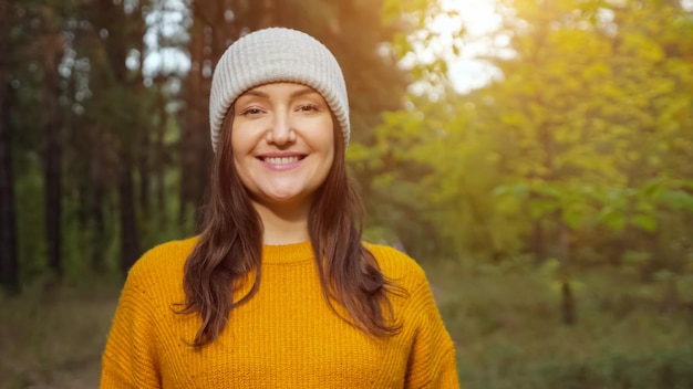 Attraktive Touristin mit warmer Strickmütze und orangefarbenem Pullover lächelt zur Kamera, die Zeit im grünen Vorstadtwald verbringt, Nahaufnahme