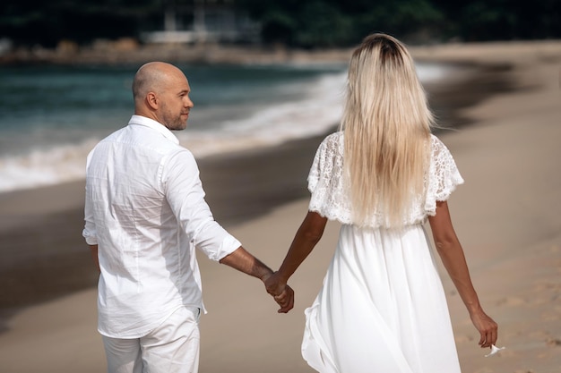 Attraktive trendige junge Paare in weißen Kleidern gehen am Strand entlang und halten sich an den Händen. Schöne Blondine mit langen Haaren und einem Glatzkopf sind im Urlaub. Phuket. Thailand. Rückansicht