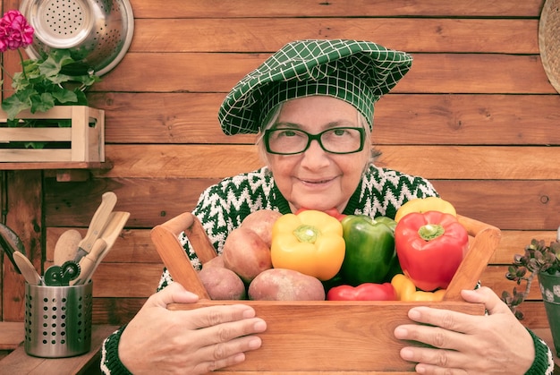 Attraktive und lächelnde ältere Frau, die einen Holzkorb voller frisch gepflückter Paprika und Kartoffeln aus dem Garten hält. Holzhintergrund