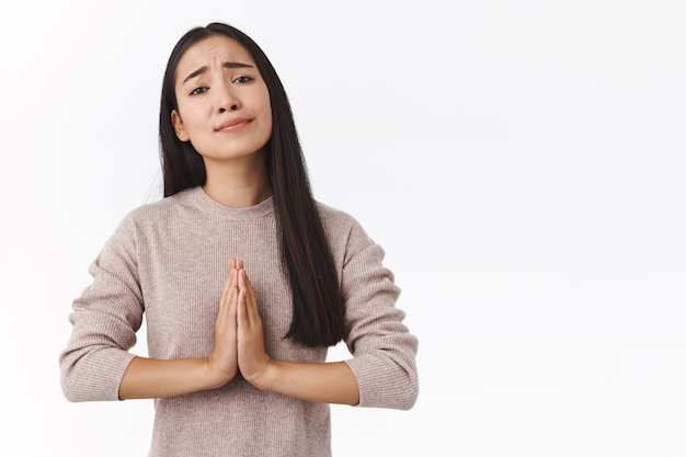 Attraktive unruhige junge ostasiatische moderne Frau mit dunklem Haar, Handflächen in der Nähe der Brust im Gebet zusammendrücken, betteln, Geld verleihen, mit Bedürftigkeit aussehen