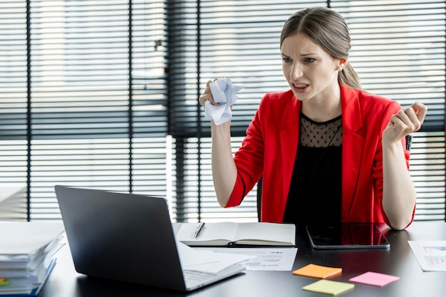 Attraktive, verrückte Geschäftsfrau sitzt wütend im Büro, zerknüllt das Papier und blickt auf den Laptop-Bildschirm