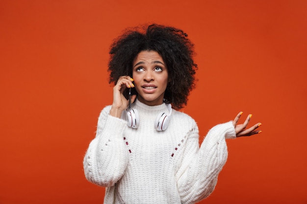 Attraktive verwirrte junge Afrikanerin mit Pullover, die auf dem Handy spricht, isoliert über roter Wand