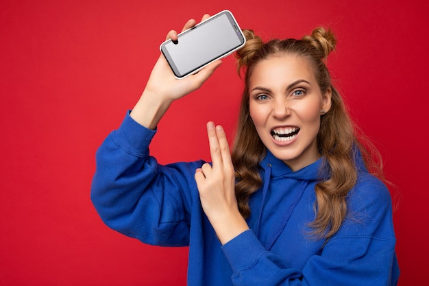 Attraktive wütende emotionale verrückte junge blonde Frau, die einen stylischen blauen Hoodie trägt, isoliert auf Rot