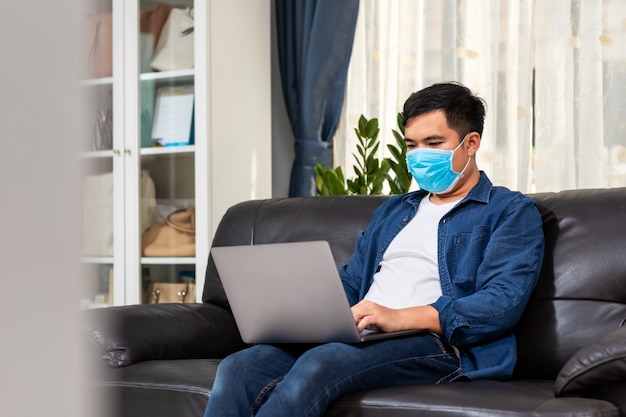 Attraktiver asiatischer junger Mann, der mit Laptop auf Couch arbeitet