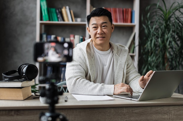 Attraktiver asiatischer Mann, der am Laptop arbeitet und Videos auf einem digitalen Smartphone aufzeichnet