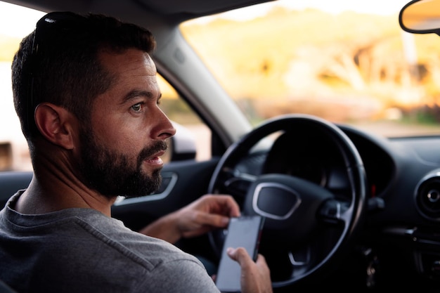 Foto attraktiver bärtiger junger mann, der sein smartphone benutzt, während er darauf wartet, in seinem auto zu sitzen freizeit und technologie des kommunikationskonzepts