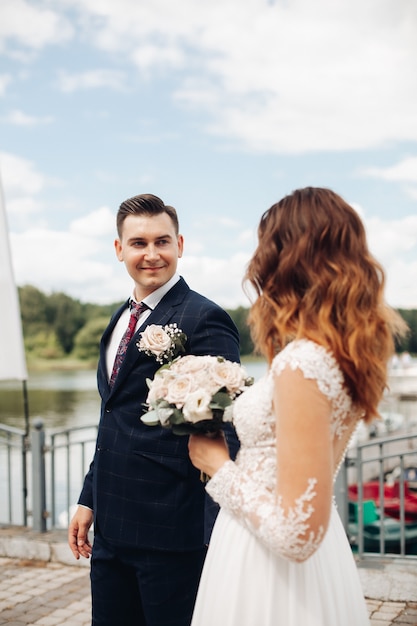 Foto attraktiver bräutigam im schwarzen anzug mit seiner schönen braut im langen weißen kleid nahe dem see