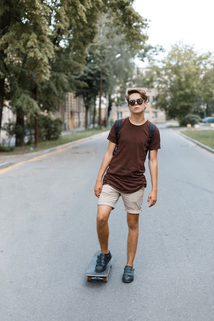 Attraktiver cooler junger Hipster-Mann mit Sonnenbrille in einem stilvollen T-Shirt in Sommershorts in Turnschuhen, die an einem warmen Sommertag auf einem Skateboard im Park reiten