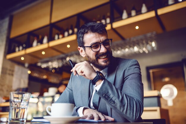 Attraktiver eleganter kaukasischer lächelnder Geschäftsmann im Anzug und mit Brillen, die im Café sitzen und in der Tagesordnung schreiben. Im Vordergrund steht Kaffee in einem Glas Wasser.