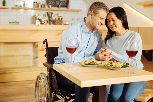 Attraktiver entzückter behinderter Mann und eine ziemlich freudige dunkelhaarige Frau, die mit geschlossenen Augen sitzt und beim romantischen Abendessen Händchen hält