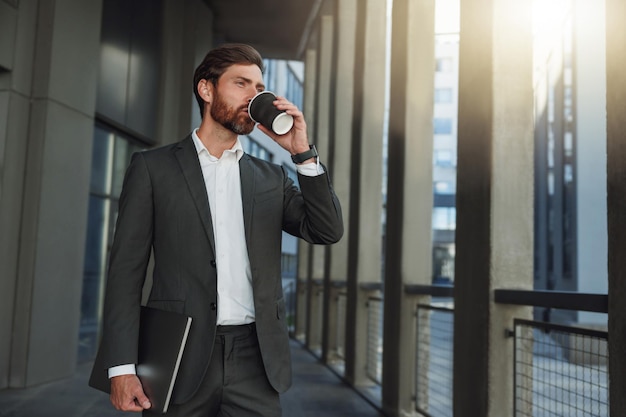 Attraktiver europäischer Geschäftsmann im Anzug, der während der Pause Kaffee in der Nähe des Bürogebäudes trinkt