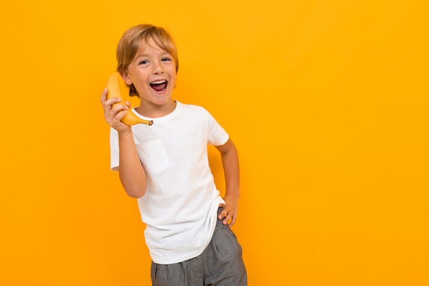 attraktiver europäischer Junge in einem weißen T-Shirt, das auf einer Banane wie am Telefon auf einer gelben Wand mit Kopienraum spricht