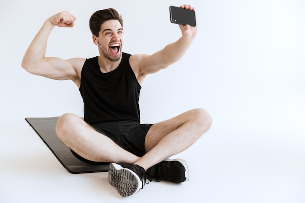 Attraktiver, fitter junger Sportler, der mit Handy auf einer Fitnessmatte sitzt, ein Selfie macht, isoliert, Muskeln spielend