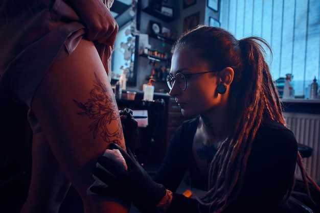 Attraktiver, fokussierter Tätowierer kreiert im dunklen Tattoo-Studio ein neues Tattoo auf dem Bein einer jungen Frau.