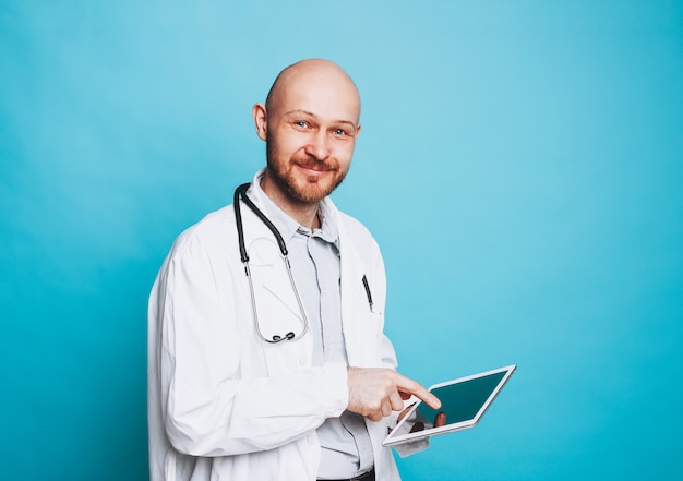 Attraktiver freundlicher kahler bärtiger lächelnder Arzt mit Tablette, die Kamera lokalisiert auf blauem Hintergrund betrachtet