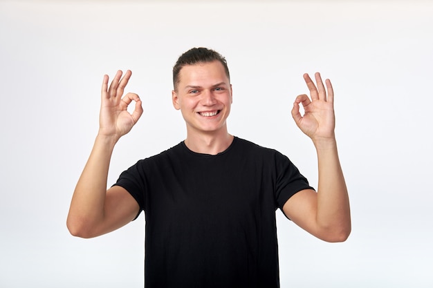 Attraktiver fröhlicher junger mann im schwarzen hemd, das ok-geste zeigt, lächelnd.
