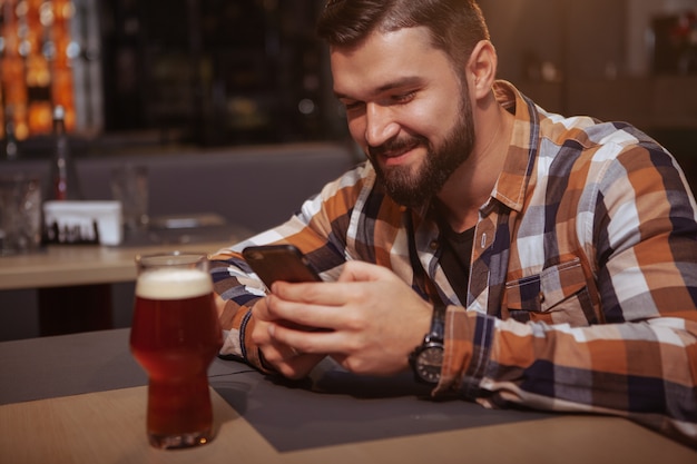 Attraktiver fröhlicher Mann, der sein Telefon benutzt, während er Bier an der Bar trinkt