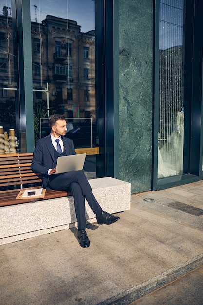 Attraktiver Geschäftsmann, der bei schönem Wetter draußen auf der Bank sitzt, während er den Laptop für die Online-Arbeit verwendet