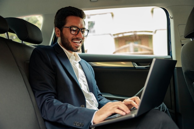 Attraktiver Geschäftsmann im Anzug arbeitet am Laptop, während er im Auto zum Büro fährt