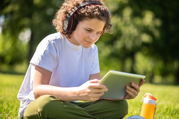 Attraktiver heranwachsender Junge in drahtlosen Kopfhörern, der mit gekreuzten Beinen auf dem Gras sitzt und auf sein Tablet schaut