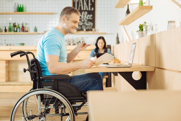 Attraktiver inspirierter behinderter Mann, der in einem Rollstuhl sitzt und ein Blatt Papier und eine Tasse Kaffee hält und an seinem Laptop arbeitet