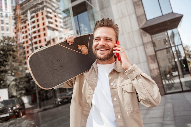 Attraktiver junger blonder lächelnder Mann, der Skateboard auf Schultern an der Stadtstraße hält, die zwischen Glasgebäuden steht. Trägt Jeans-Outfit. Sportlich stylischer Skateboarder spricht über Handy-Lauphing.