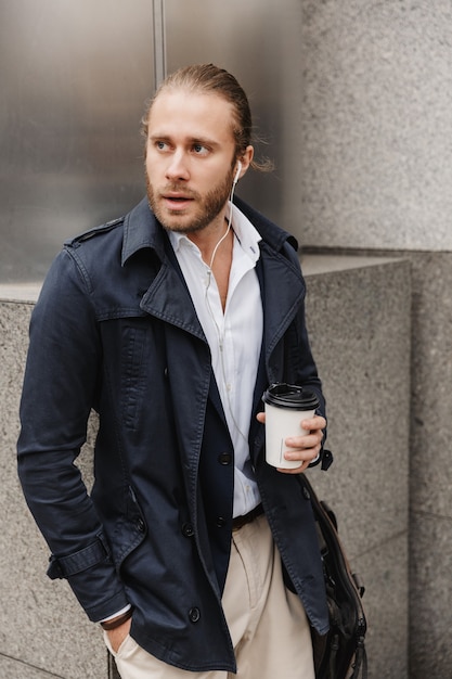 Attraktiver junger blonder Mann in formeller Kleidung, der über Kopfhörer spricht und eine Kaffeetasse zum Mitnehmen hält, während er im Freien auf einer Stadtstraße steht