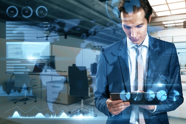 Attraktiver junger europäischer Geschäftsmann mit Tablet mit abstrakter Karte und Hologramm für Geschäftsdiagramme auf verschwommenem Hintergrund im Bürointerieur Investorenfinanzierung und Finanz-Dashboard-Konzept Doppelbelichtung