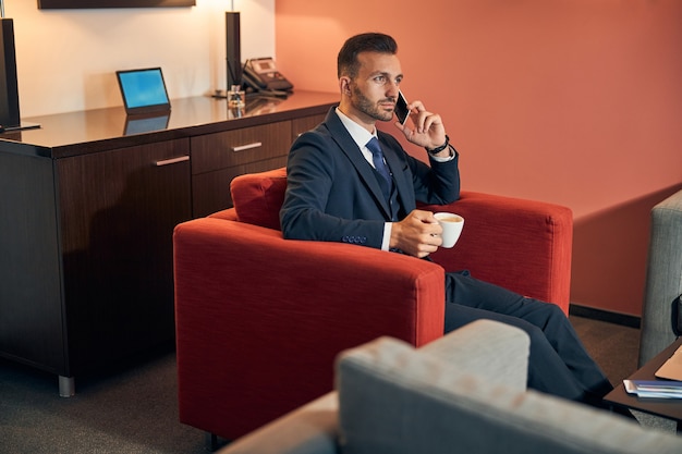 Attraktiver junger Geschäftsmann, der mit einer Tasse Kaffee im Sessel sitzt und während des Telefongesprächs konzentriert aussieht