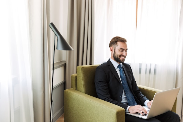 Attraktiver junger Geschäftsmann im Anzug, der auf einem Stuhl im Hotelzimmer sitzt und an einem Laptop arbeitet?