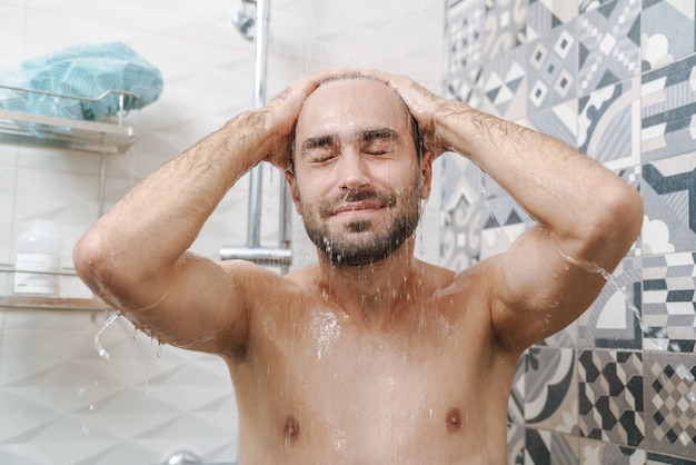 Attraktiver junger lächelnder Mann, der Haare wäscht, während er in der Dusche steht