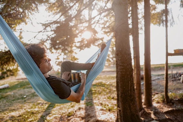 Attraktiver junger Mann, der sich in der Hängematte mit Kaffeetee in der Hängematte im hinteren Wald ausruht