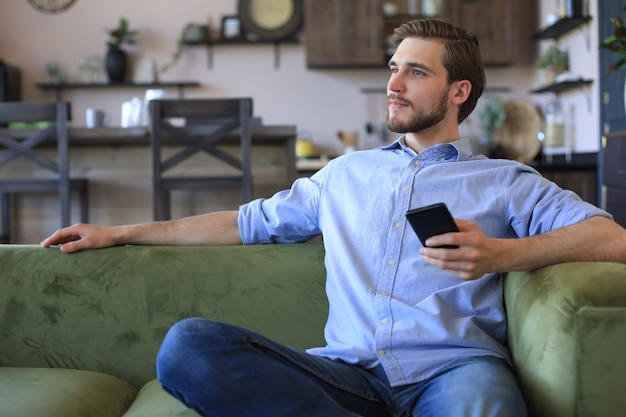 Attraktiver junger Mann, der sich zu Hause auf einer Couch entspannt und mit dem Handy soziale Netze überprüft.