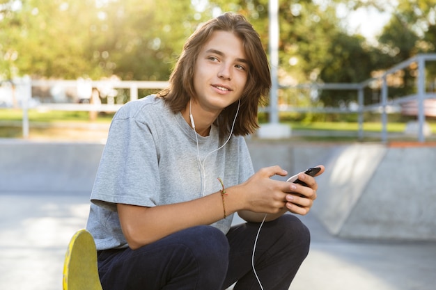 Attraktiver junger Mann, der Zeit im Skatepark verbringt und Musik mit Kopfhörern hört