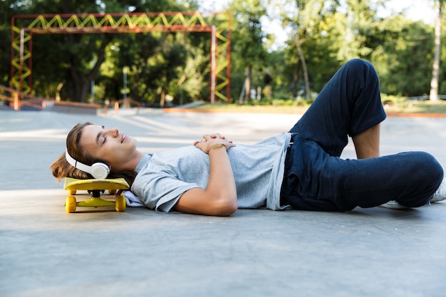 Attraktiver junger Mann, der Zeit im Skatepark verbringt und Musik mit Kopfhörern hört