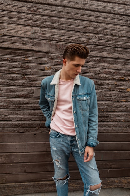 Attraktiver junger Mann mit Frisur in stylischem rosa T-Shirt in modischer Jeansjacke posiert in der Nähe der braunen Vintage-Holzwand in der Straße. Europäisches gutaussehendes Kerlmodell in Jeanskleidung steht in der Stadt.