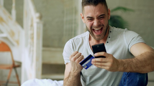 Attraktiver junger Mann mit Smartphone- und Kreditkarteneinkäufen im Internet sitzt zu Hause auf dem Bett