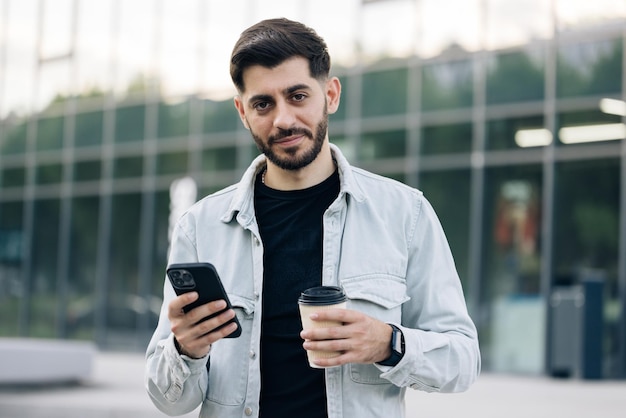 Attraktiver kaukasischer armenischer junger bärtiger mann geht die straße entlang und benutzt telefonlächeln sonnenlicht