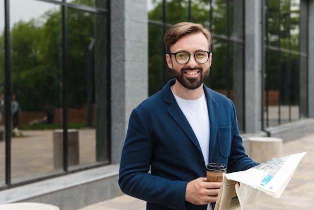 Attraktiver lächelnder junger bärtiger Mann mit Jacke, der Zeitung liest, während er im Freien in der Stadt steht und Kaffee zum Mitnehmen trinkt