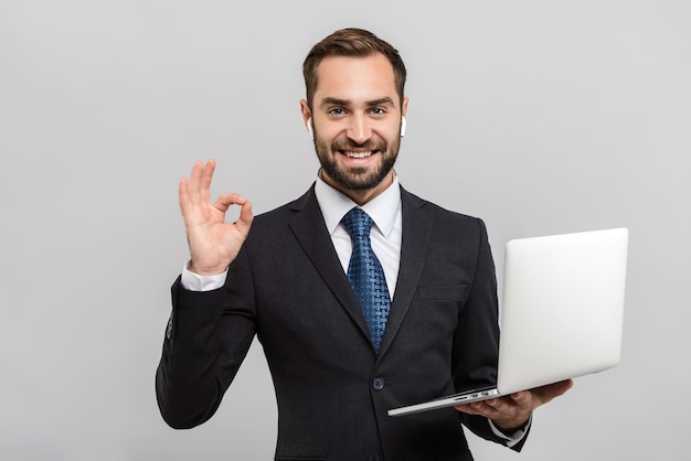 Attraktiver lächelnder junger Geschäftsmann im Anzug, der isoliert über grauer Wand steht, mit Laptop-Computer, ok Geste