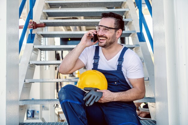 Attraktiver lächelnder kaukasischer Arbeiter in Overalls, die auf Treppen sitzen und auf dem Smartphone sprechen. Raffinerie außen.