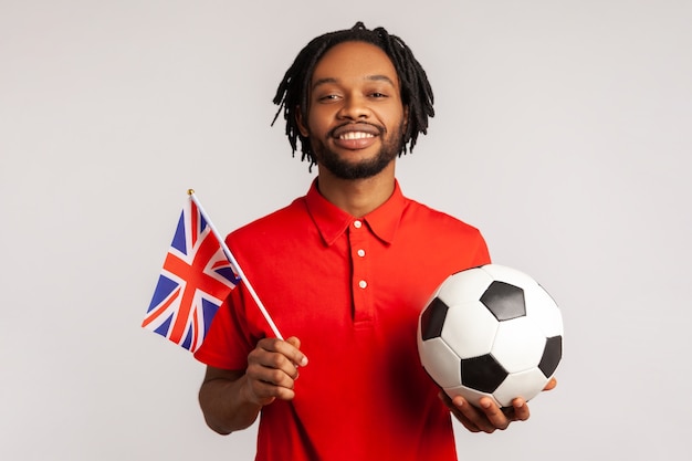Attraktiver lächelnder Mann, der britische Flagge und Fußballschwarzweiss-Ball hält, vereinigte Fußballliga.