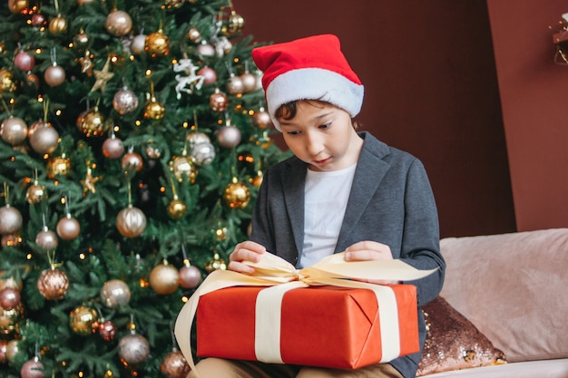 Attraktiver lustiger Tweenjunge mit dem dunklen Haar in der offenen Geschenkbox Sankt-Hutes auf Weihnachtsbaum